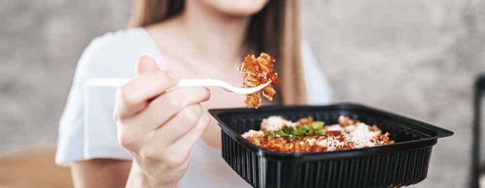 Saboroso plato de comida italiana en un recipiente negro, con pasta, queso y salsa, sostenido por una persona con camiseta blanca, en un entorno informal y relajado.